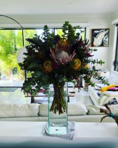 An arrangement of wild and native flowers in a glass vase, with a white leather couch suite in the background