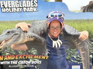 The smiling face of a woman wearing glasses, posing through a billboard of a man holding an alligator over his shoulders, Everglades Park, Florida, USA