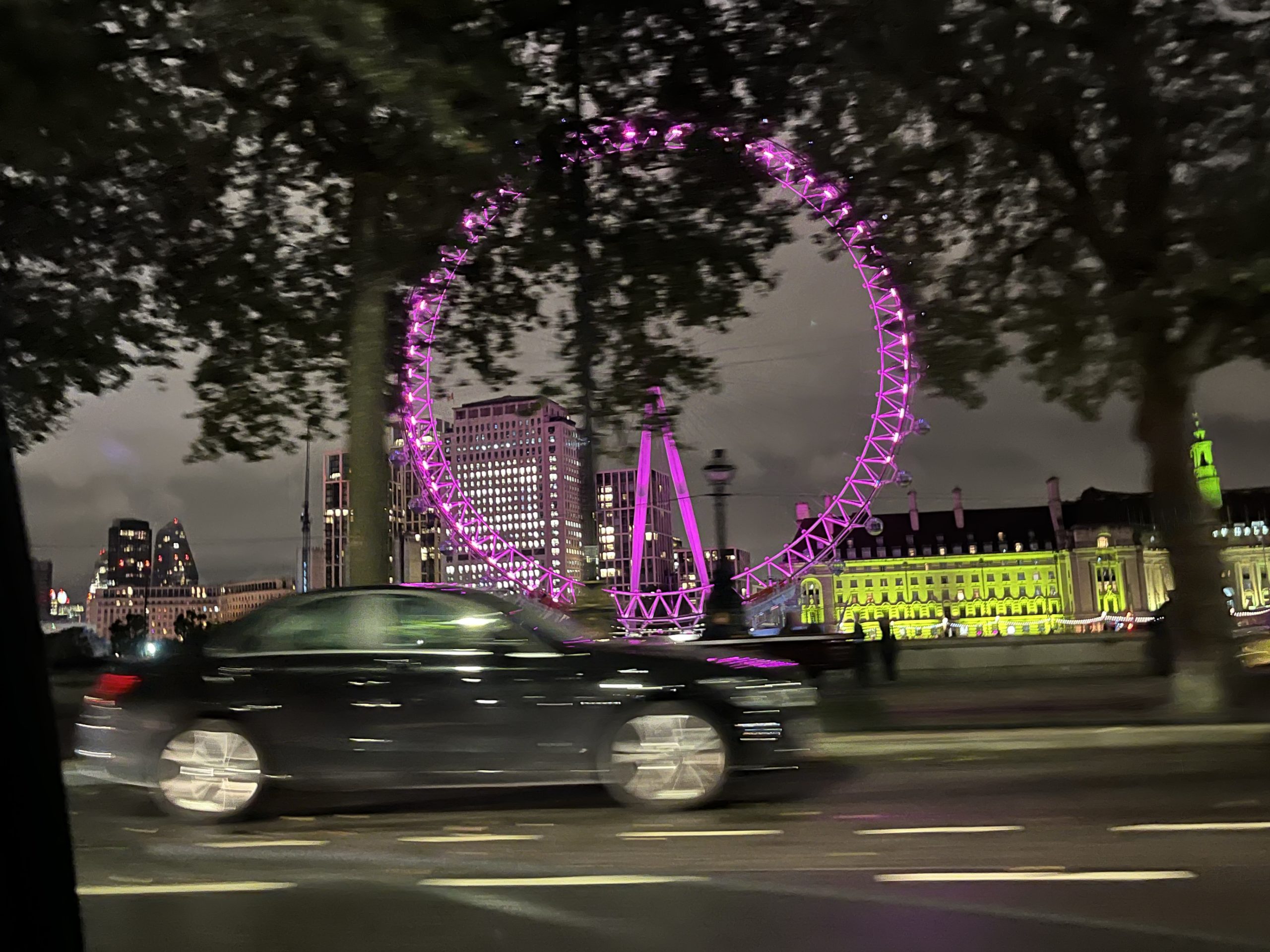 Night time shot of a black car in motion on a road, in the background some buildings and a ferris wheel lit in bright pink lights